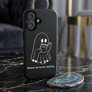 Peut inclure: Coque de téléphone noire avec une illustration blanche d'un fantôme lisant un livre et le texte "Rather be home reading". La coque est posée sur une surface en marbre noir avec une tranche d'agate bleue et grise.