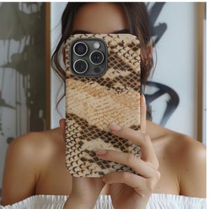 Peut inclure: Une coque de smartphone avec un motif peau de serpent dans des tons marron et beige. L'étui a un module caméra noir avec trois lentilles. Le téléphone est tenu devant une personne.