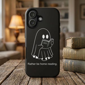 Peut inclure: Coque de téléphone noire avec une illustration blanche d'un fantôme lisant un livre. Le texte "Rather be home reading" est en dessous du fantôme. La coque est sur une surface en bois.