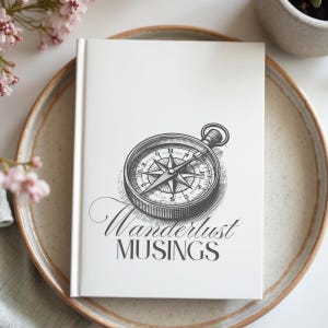 May include: A white journal with a detailed compass illustration and the words "Wanderlust Musings" in elegant script. The journal is on a ceramic plate, with pink flowers in the background.