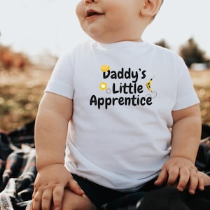 May include: A white short-sleeved t-shirt featuring the text "Daddy's Little Apprentice" in black, with yellow and black graphics of a hard hat, gear, and drill. The shirt is worn by a baby.