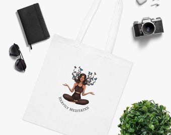 May include: White tote bag featuring an illustration of a person meditating with butterflies around their head. The text "HEAVILY MEDITATED" is below the illustration. Other items in the image include a black notebook, sunglasses, and a camera.