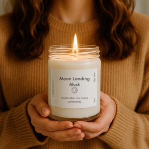 May include: A lit soy wax candle in a glass jar, labeled "Moon Landing Musk." The label includes a graphic and the text "Smells fake... but pretty convincing." The candle is held in both hands.