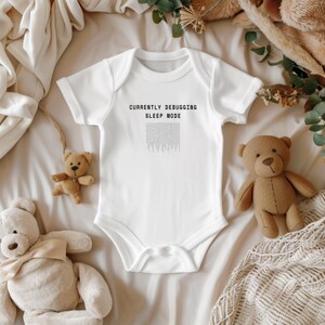 Puede incluir: Body de bebé blanco con el texto negro "CURRENTLY DEBUGGING SLEEP MODE". La imagen incluye ositos de peluche y elementos decorativos sobre una manta color crema.