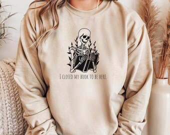 May include: A beige sweatshirt with a black skeleton illustration reading a book. The text on the sweatshirt reads "I closed my book to be here."