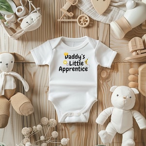 Op de afbeelding: Witte babybody met de tekst "Daddy's Little Apprentice" in zwart, geel en lichtblauw. De body is omringd door babyspeelgoed, waaronder knuffels en houten blokken, op een lichtbruin houten oppervlak.