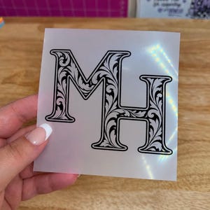 May include: A square, semi-transparent transfer sheet featuring the black capital letters M and H. The letters are designed with an intricate, swirling floral filigree pattern throughout their interior, enclosed by a bold, solid black outline.