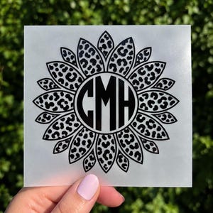 May include: A black vinyl decal featuring a sunflower design with leopard print patterns on the petals and a central circular monogram containing the letters CMH. The decal is presented on a square white backing sheet with a smooth finish.