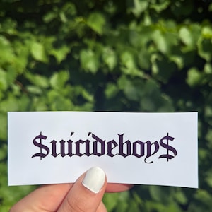 May include: A rectangular white sticker featuring the text $uicideboy$ printed in a bold, black, gothic-style font. The sticker is presented against a natural outdoor background of green leaves, showcasing the clear, dark lettering on the white surface.