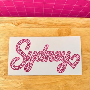 May include: A white rectangular decal featuring the name Sydney written in a cursive script font. The lettering is filled with a dark pink leopard print pattern and ends with a small heart shape integrated into the final letter of the name.