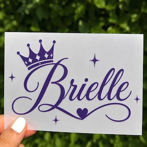 May include: A white rectangular decal featuring the name Brielle in a purple cursive script font. The design includes a purple crown icon above the name, a small purple heart at the end of the lettering, and four purple four-pointed star accents.