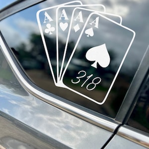 May include: White decal of four playing card aces on a car window. The suits are clubs, hearts, diamonds, and spades. The number "318" is below the spade card. The car is gray.