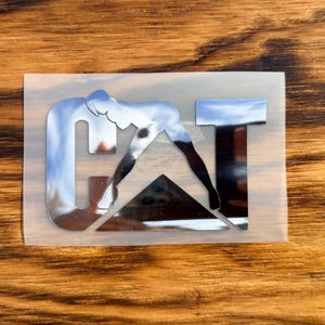 Puede incluir: Una pegatina cromada con una figura estilizada que se inclina para tocarse los dedos de los pies, formando la letra "G". Las letras "AT" tambi&eacute;n son cromadas. La pegatina est&aacute; sobre un respaldo transparente.