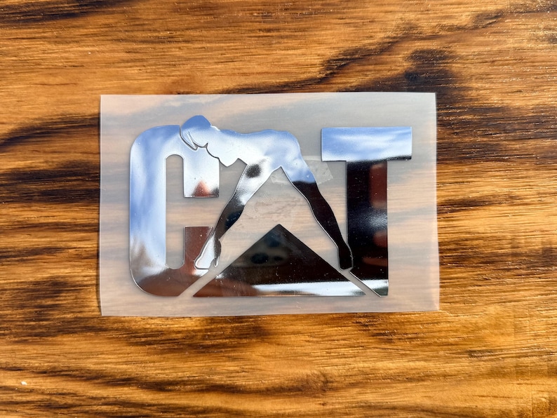 Puede incluir: Una pegatina cromada con una figura estilizada que se inclina para tocarse los dedos de los pies, formando la letra "G". Las letras "AT" tambi&eacute;n son cromadas. La pegatina est&aacute; sobre un respaldo transparente.