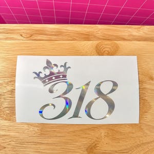 May include: A rectangular white decal featuring a holographic crown graphic positioned above the number 318 in a large, elegant, cursive-style font. The design reflects light with a multi-colored sheen, creating a metallic appearance on the surface.