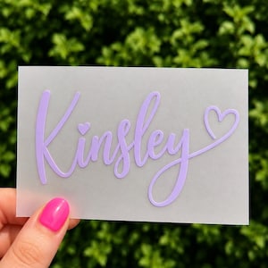 May include: A rectangular, semi-transparent frosted sheet featuring the name Kinsley written in a light purple, cursive script font. A small heart is positioned above the letter i, and a larger heart follows the final letter y.