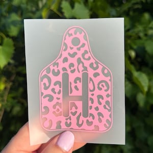 May include: A pink livestock ear tag decal featuring a grey leopard print pattern and the letter H in the center. The decal is printed on a translucent backing sheet, showing a cutout hole at the top for attachment purposes.