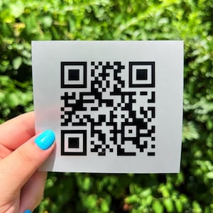 May include: A square white sticker featuring a black QR code pattern printed in the center. The design consists of a standard matrix of black squares and pixels arranged to form a scannable digital code on a smooth, flat, white surface.