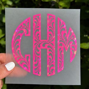 May include: A circular monogram decal featuring the letters C, H, and M in a bright neon pink color with a holographic finish. The letters are stylized with intricate, swirling filigree cutouts and are applied to a translucent backing sheet.