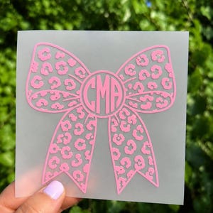 May include: A pink vinyl decal featuring a bow shape with a leopard print pattern and a central circular monogram containing the letters CMA. The design is presented on a translucent backing sheet, showcasing a clear and detailed graphic.