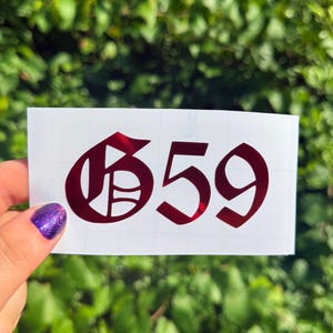 May include: A rectangular white adhesive transfer sheet featuring the number 659 in a dark red, metallic, gothic-style font. The numbers are centered on the backing paper, which has a faint grid pattern visible across the surface.