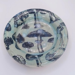 May include: A small, round ceramic dish with a light blue glaze. The dish is decorated with a repeating pattern of dark blue mushroom illustrations. The rim of the dish is slightly raised.
