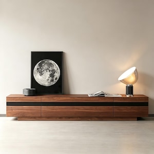 May include: A modern, low-profile wooden media console with a dark wood finish and black accent stripes. A framed black and white moon print and a stylish lamp with a black base and white shade sit atop the console. A stack of black plates and an open book are also visible.
