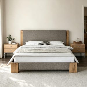Puede incluir: Una cama moderna con cabecero tapizado gris y estructura de madera. La cama está hecha con ropa de cama blanca, una manta gris y una almohada con estampado en blanco y negro. Dos mesitas de noche de madera con cajones flanquean la cama.