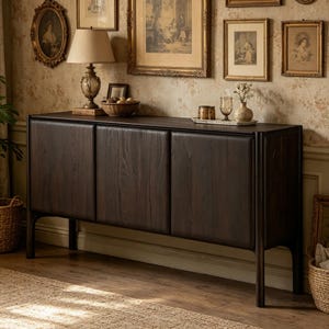Puede incluir: Aparador de madera marrón oscuro con tres puertas y cuatro patas. El mueble está en una habitación con obras de arte enmarcadas y una lámpara. El aparador es de madera y tiene un patrón de vetas natural. La habitación tiene una alfombra y una cesta.