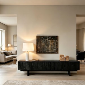 May include: A modern living room featuring a black wooden console table with a textured front, a white lamp, and abstract artwork. The room has neutral tones, a rug, and a sofa in the background, creating a minimalist aesthetic.