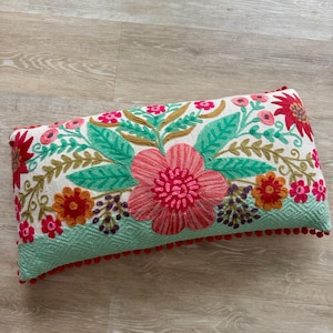 May include: A white pillow with a green and blue background and a floral embroidery design in shades of pink, red, orange, and yellow. The pillow has a red pom-pom trim.