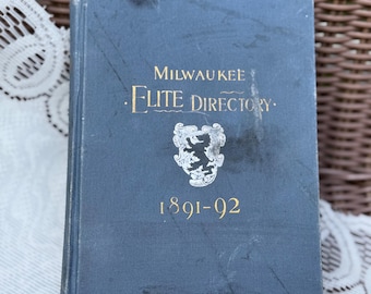 Op de afbeelding: Een vintage blauw boek met de titel "Milwaukee Elite Directory" uit 1891-92. De omslag heeft gouden letters en een wapen. Het boek is licht versleten, wat duidt op de leeftijd.