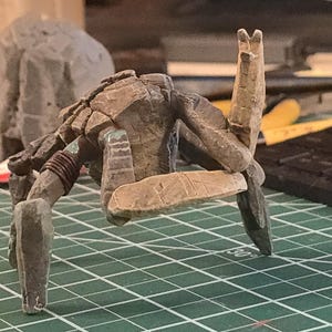 May include: A small, multi-legged creature figurine with a textured, weathered brown and gray finish. The figure features a segmented shell-like back, a raised limb, and a flat, rectangular base component held between its front appendages.