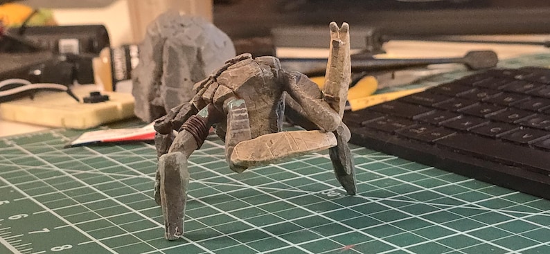 May include: A small, multi-legged creature figurine with a textured, weathered brown and gray finish. The figure features a segmented shell-like back, a raised limb, and a flat, rectangular base component held between its front appendages.
