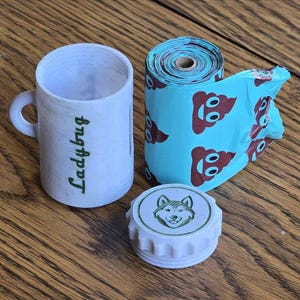Op de afbeelding: Een kleine witte mok met het woord "Ladybug" in groen schrift, een rol blauwe hondenpoepzakjes met bruine poep-emoji's en een wit deksel met een wolf-illustratie.