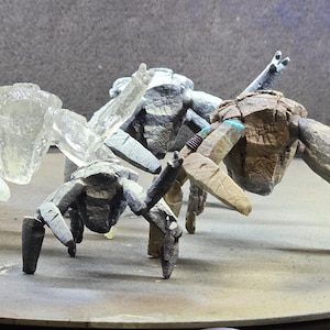 May include: Three multi-legged crab-like figurines standing in a group. One figure is translucent and clear, while the other two are opaque with textured, weathered surfaces in shades of gray and brown, featuring articulated limbs and segmented bodies.