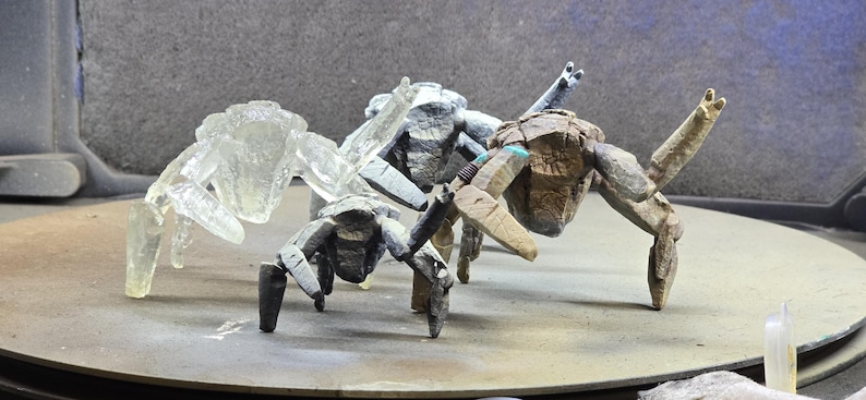 May include: Three multi-legged crab-like figurines standing in a group. One figure is translucent and clear, while the other two are opaque with textured, weathered surfaces in shades of gray and brown, featuring articulated limbs and segmented bodies.