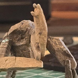 May include: A miniature stone-like creature figurine with a textured, rocky surface. The figure has a central upright torso, multiple angular limbs, and etched geometric markings on its surface. One limb is wrapped with a dark brown coiled material.
