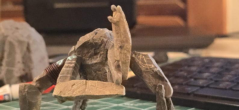 May include: A miniature stone-like creature figurine with a textured, rocky surface. The figure has a central upright torso, multiple angular limbs, and etched geometric markings on its surface. One limb is wrapped with a dark brown coiled material.