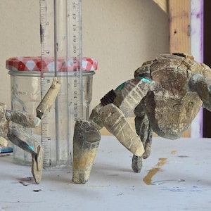 May include: Two articulated crab-like figures crafted from textured, stone-colored material with small teal accents. A clear glass jar with a red and white checkered lid stands between them, positioned next to a ruler showing a height of 4 inches.