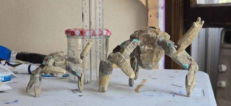May include: Two articulated crab-like figures crafted from textured, stone-colored material with small teal accents. A clear glass jar with a red and white checkered lid stands between them, positioned next to a ruler showing a height of 4 inches.