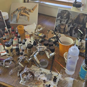 May include: A cluttered artist workspace featuring multiple small, articulated creature figurines in various stages of assembly, numerous paint bottles, brushes in glass jars, water cups, and printed reference images of the models.