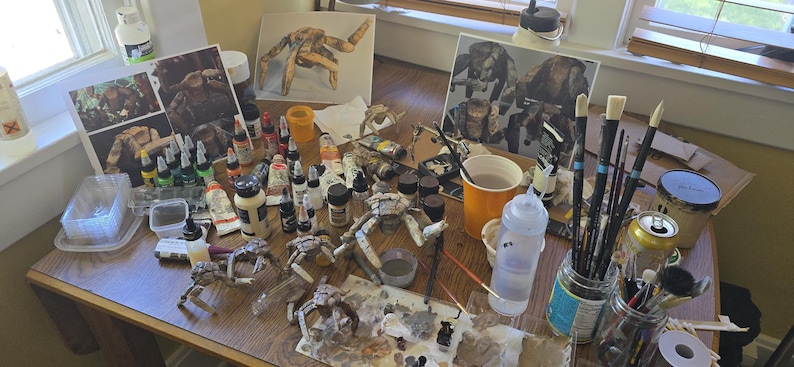May include: A cluttered artist workspace featuring multiple small, articulated creature figurines in various stages of assembly, numerous paint bottles, brushes in glass jars, water cups, and printed reference images of the models.