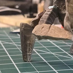 May include: A brown, multi-legged creature figurine with a textured, rock-like surface and carved markings on its limbs. The figure stands on a green cutting mat with white grid lines, showing a portion of the creature's legs and torso structure.