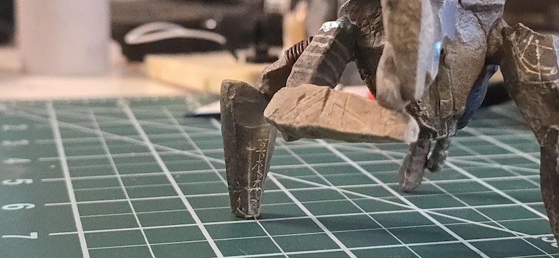 May include: A brown, multi-legged creature figurine with a textured, rock-like surface and carved markings on its limbs. The figure stands on a green cutting mat with white grid lines, showing a portion of the creature's legs and torso structure.