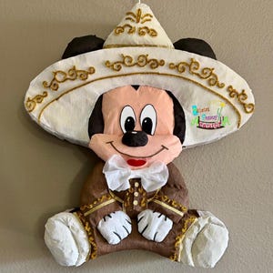 May include: A Mickey Mouse pi&ntilde;ata wearing a sombrero and traditional Mexican clothing. The pi&ntilde;ata is mainly brown, white, and gold, with a white bow tie and a smiling face. The sombrero has gold glitter detailing.