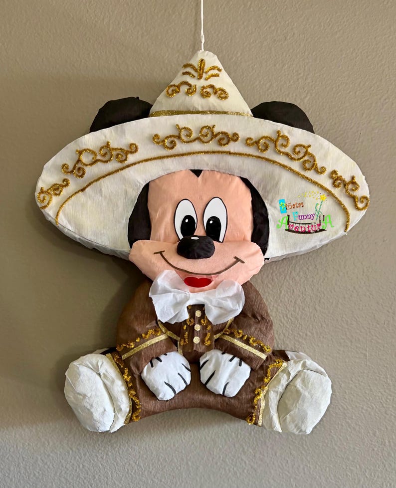 May include: A Mickey Mouse pi&ntilde;ata wearing a sombrero and traditional Mexican clothing. The pi&ntilde;ata is mainly brown, white, and gold, with a white bow tie and a smiling face. The sombrero has gold glitter detailing.