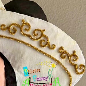 May include: A white pi&ntilde;ata with gold glitter detailing and the text "Pi&ntilde;atas Funny Adventures". The pi&ntilde;ata features black and pink elements. Gold accents are in a decorative swirl pattern.