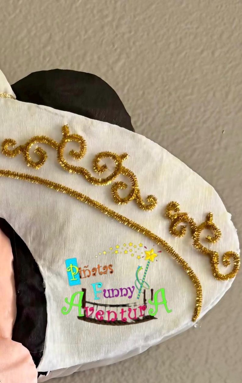 May include: A white pi&ntilde;ata with gold glitter detailing and the text "Pi&ntilde;atas Funny Adventures". The pi&ntilde;ata features black and pink elements. Gold accents are in a decorative swirl pattern.