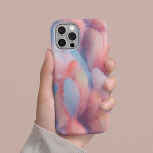 May include: A smartphone case featuring a colorful feather design in shades of pink, blue, and peach. The case has a smooth finish and a camera cutout. The phone case is held by a person.